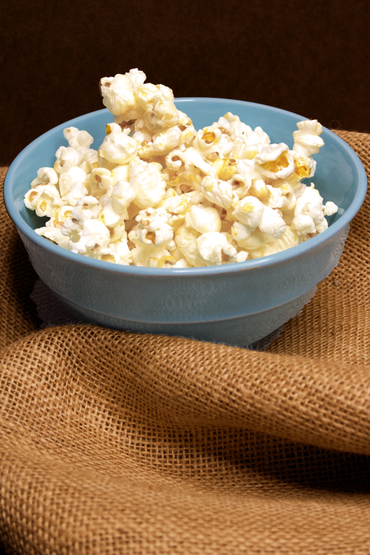 A blue bowl piled high with fresh Homemade Kettle Corn sits atop a rumpled piece of burlap.
