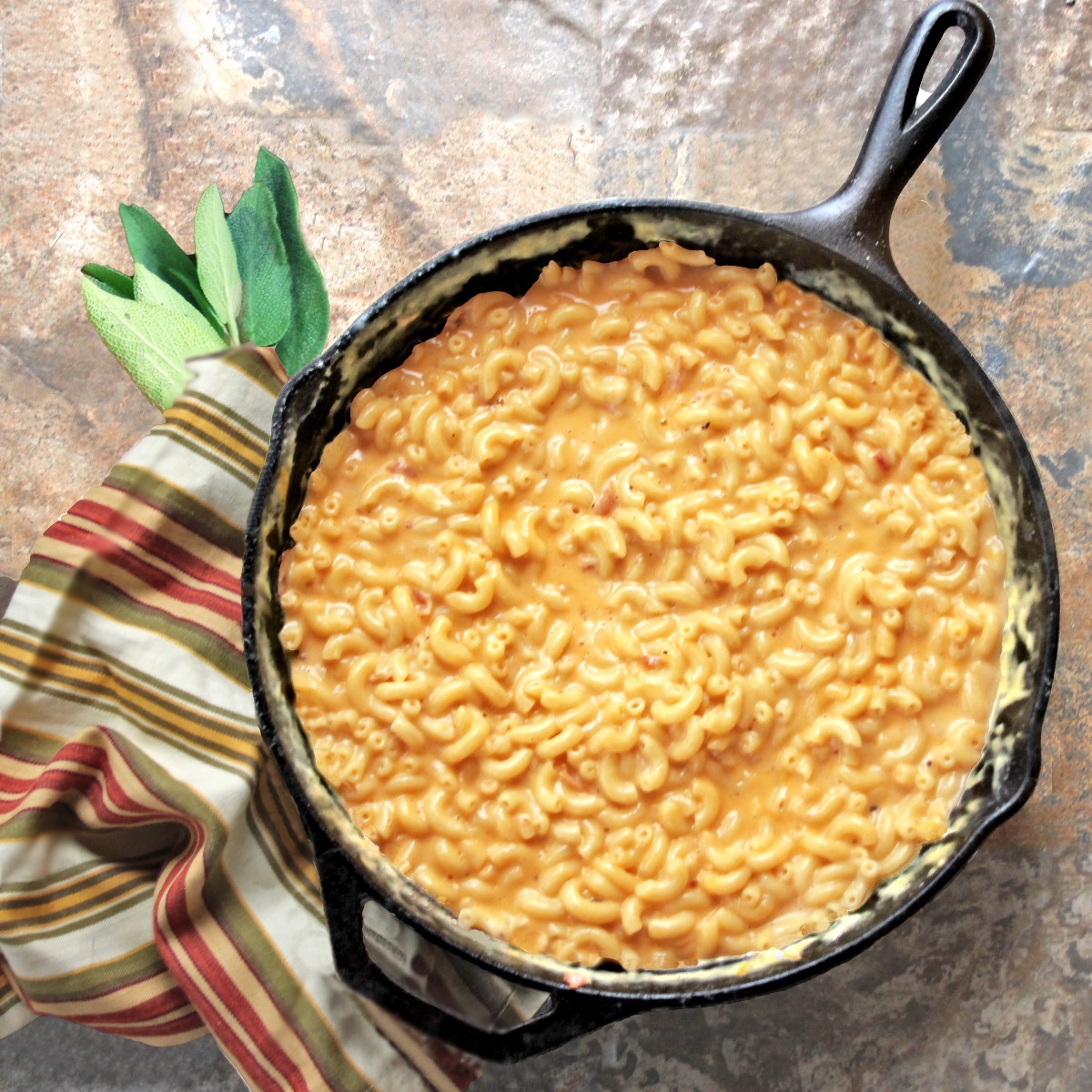 Creamy One-Pot Macaroni and Cheese in a cast-iron skillet, next to a striped cloth and a bunch of sage. This macaroni and cheese is creamy and decadent, but without a roux or actual cream. Just loads of cheesy flavor!