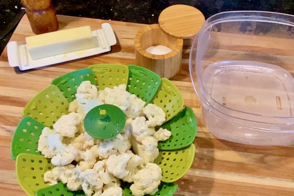 Top view of microwaved cauliflower inside a rubber colander next to a plastic bowl with salt, pepper and butter all on a cutting board. 