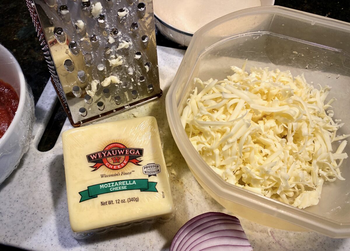 top view of a block of mozzarella, a cheese grater and a bowl of freshly shredded mozzarella.