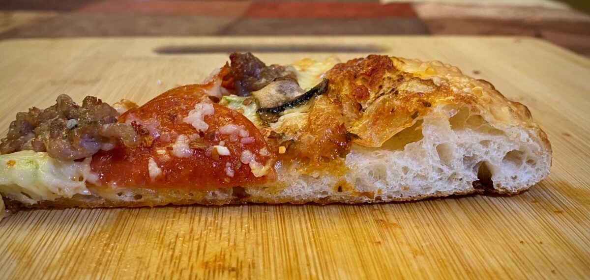 Close up side view of the airy crust in a slice of pepperoni and sausage pizza on a wooden cutting board.