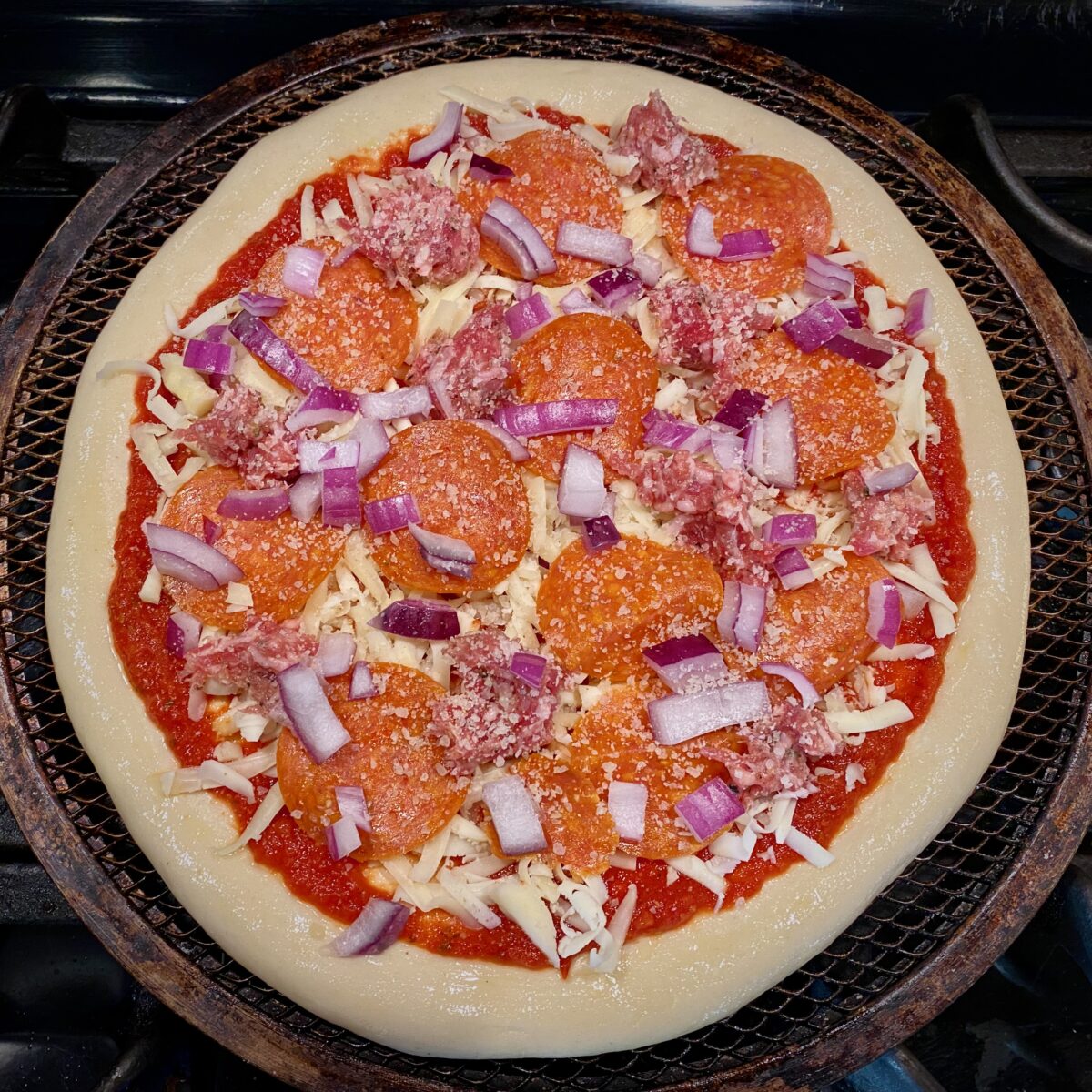 top view of a pizza crust that is topped with pizza sauce, cheese, pepperoni, sausage and. red onion.