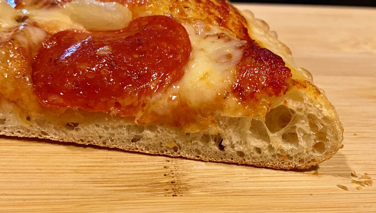 Close up side view of the airy crust of a slice of pepperoni and onion pizza.