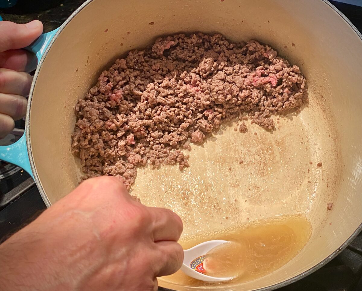 Top view of 45 degree tilted pot of browned ground beef. The beef is positioned to one side allowing the fat to drip into the bottom. A small spoon is scooping out and removing the fat.
