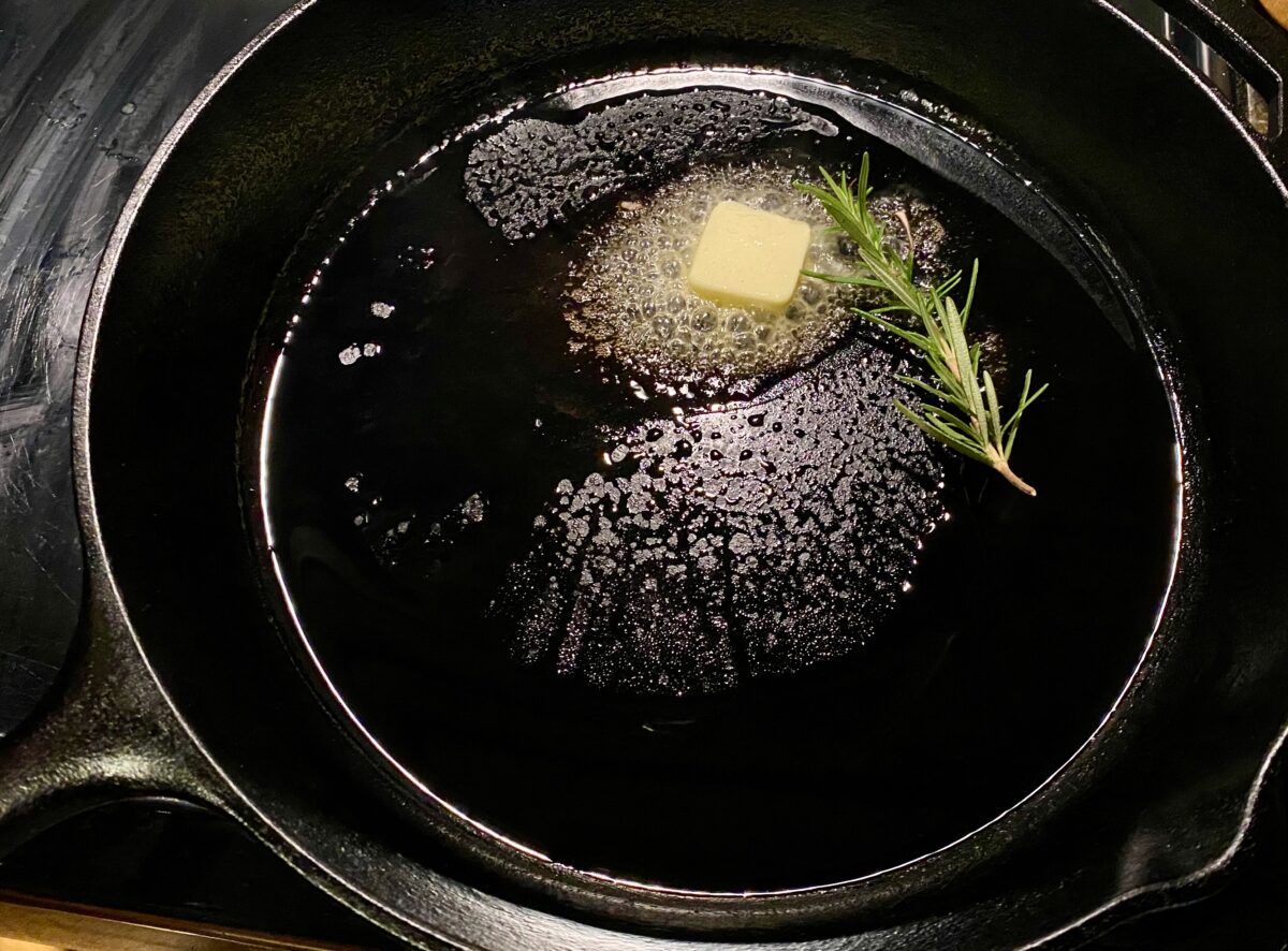Top view of a hot cast iron skillet with a tablespoon of butter and oil with a sprig of fresh rosemary.