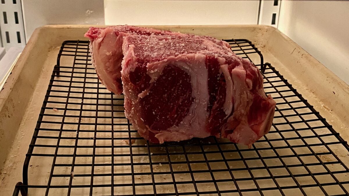 Salted thick ribeye resting in a refrigerator on a cooling rack.