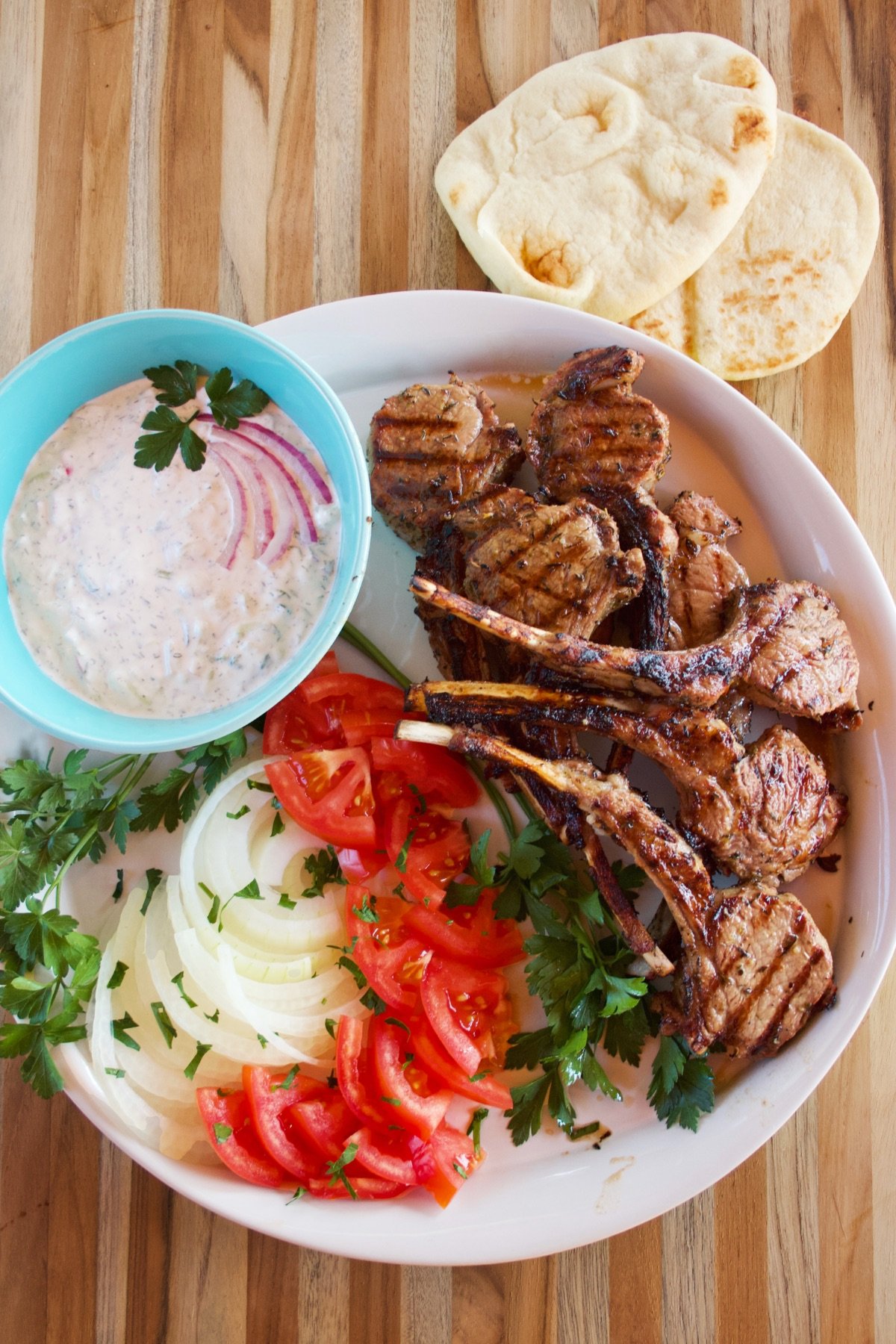 A platter loaded up with Greek Marinated Lamb chops, sliced tomatoes and onions, parsley, and a bowl of tzatziki sauce. Two pieces of naan bread sit nearby.