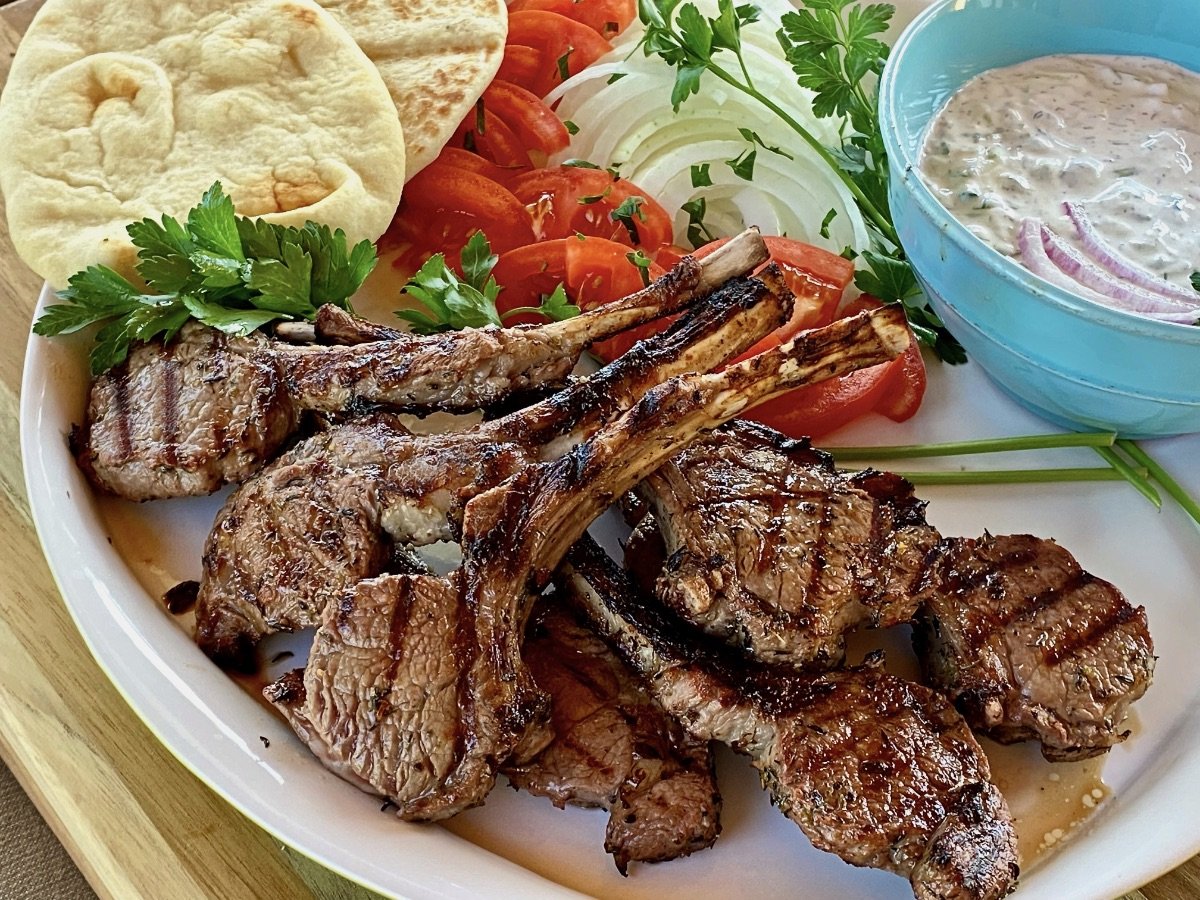 Finished Greek Marinated Lamb Chops on a platter with a bowl of tzatziki sauce, parsley sprig, naan bread, and sliced onions and tomatoes.