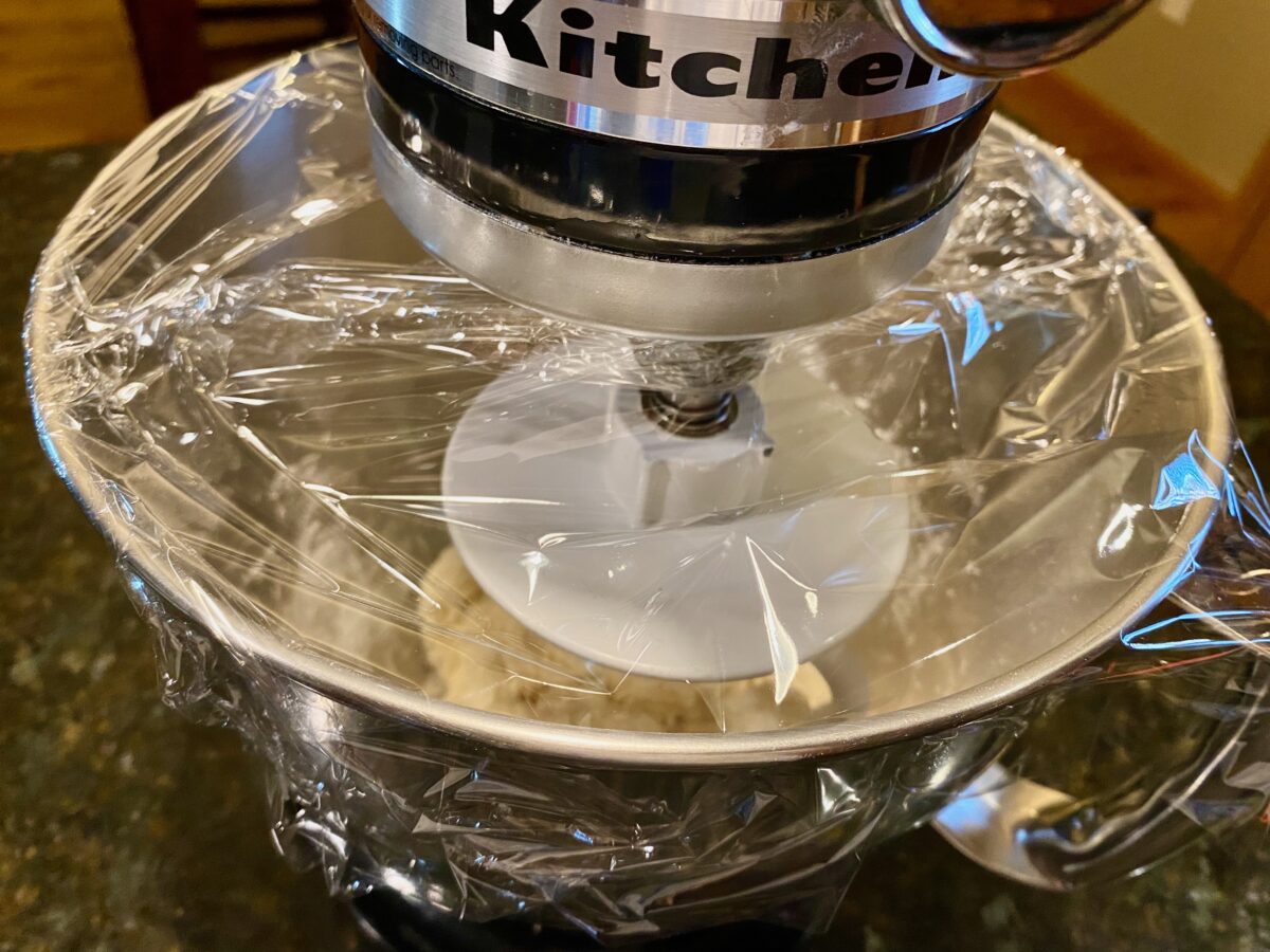 The shaggy dough is resting in the mixing bowl of the stand mixer, covered with plastic wrap.