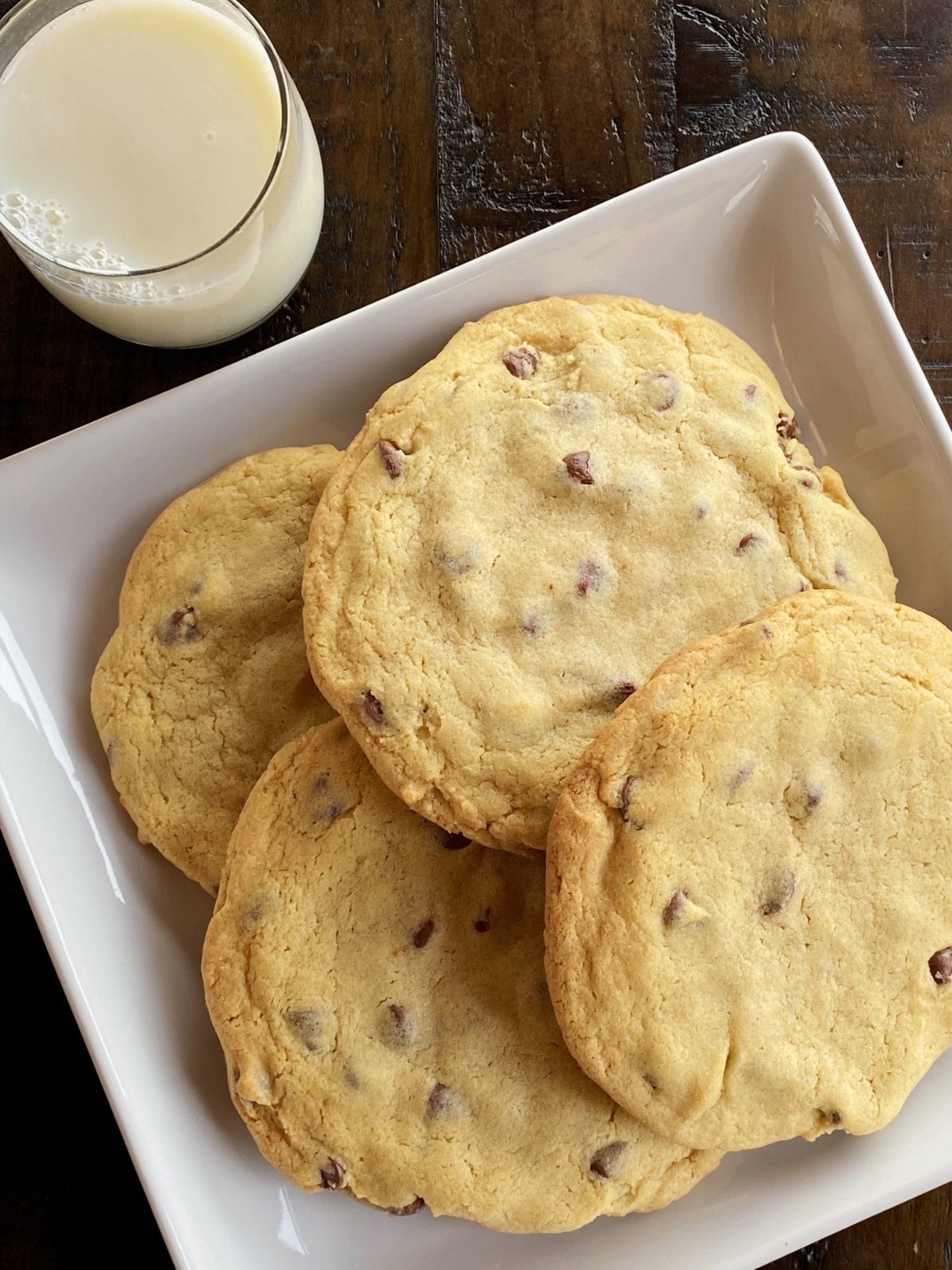 Four monster chocolate chip cookies on a square, white plate next to a glass of milk.