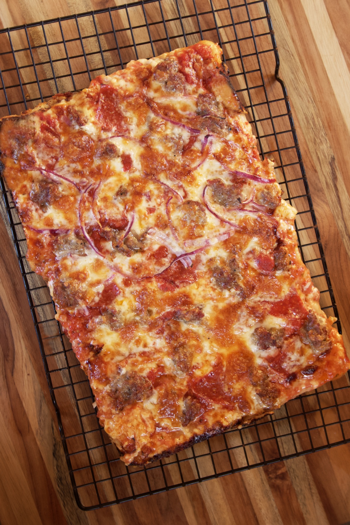 A homemade pan pizza topped with sausage, pepperoni, and onions, cooling on a wire rack.