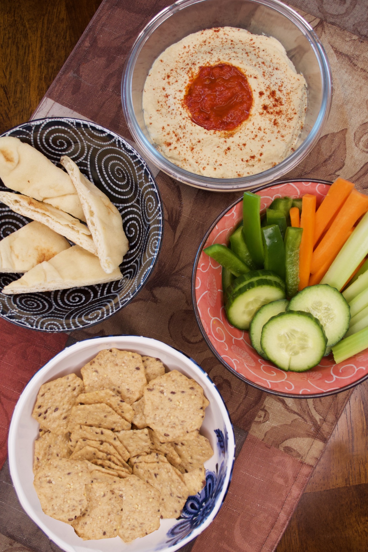 A bowl of Easy Spicy Hummus with bowls of some of our favorite snacks for dipping, including sliced vegetables (celery, carrots, cucumber, and green pepper), crackers, and pita.