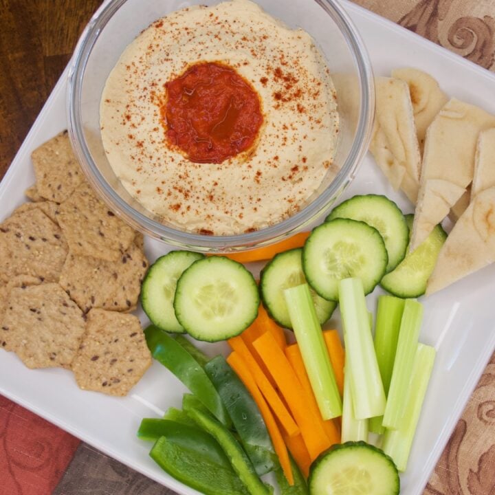 A top view of a platter with a bowl of the spicy hummus served with whole-grain crackers, naan wedges, and sliced cucumbers, carrots, and green peppers.