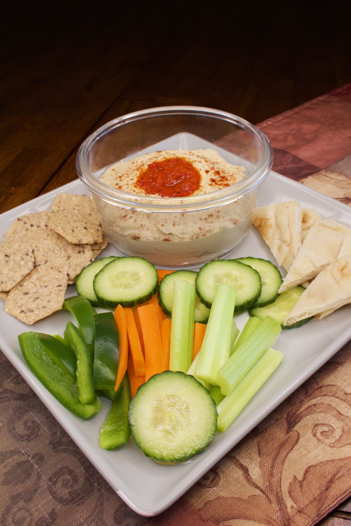 Easy Spicy Hummus on a platter with whole-grain crackers, naan wedges, and sliced cucumbers, celery, and carrots. In addition to the harissa topping, we've also sprinkled the hummus with a Berbere spice blend.