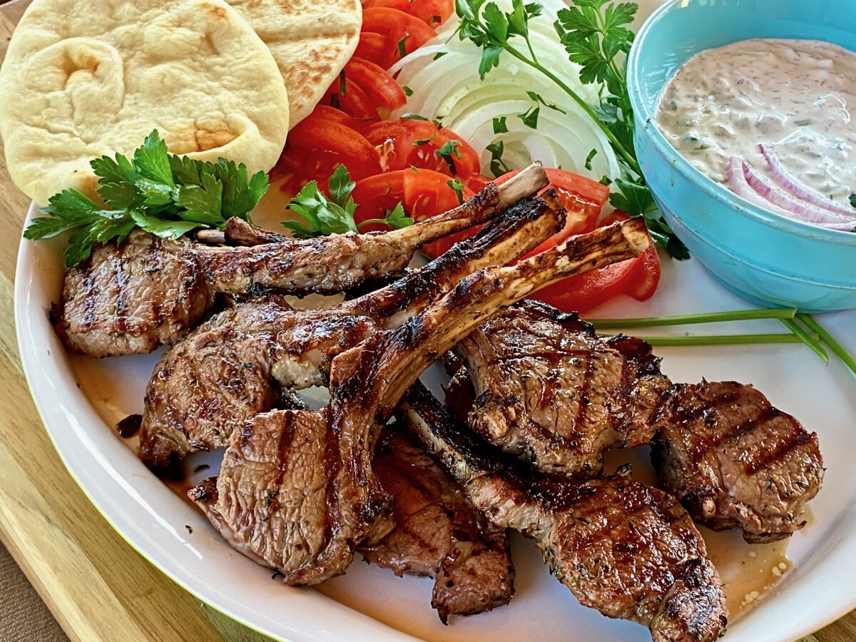 Plate of grilled greek marinated lamb chops with sliced onions and tomatoes alongside pita bread and a bowl of tzatziki sauce.