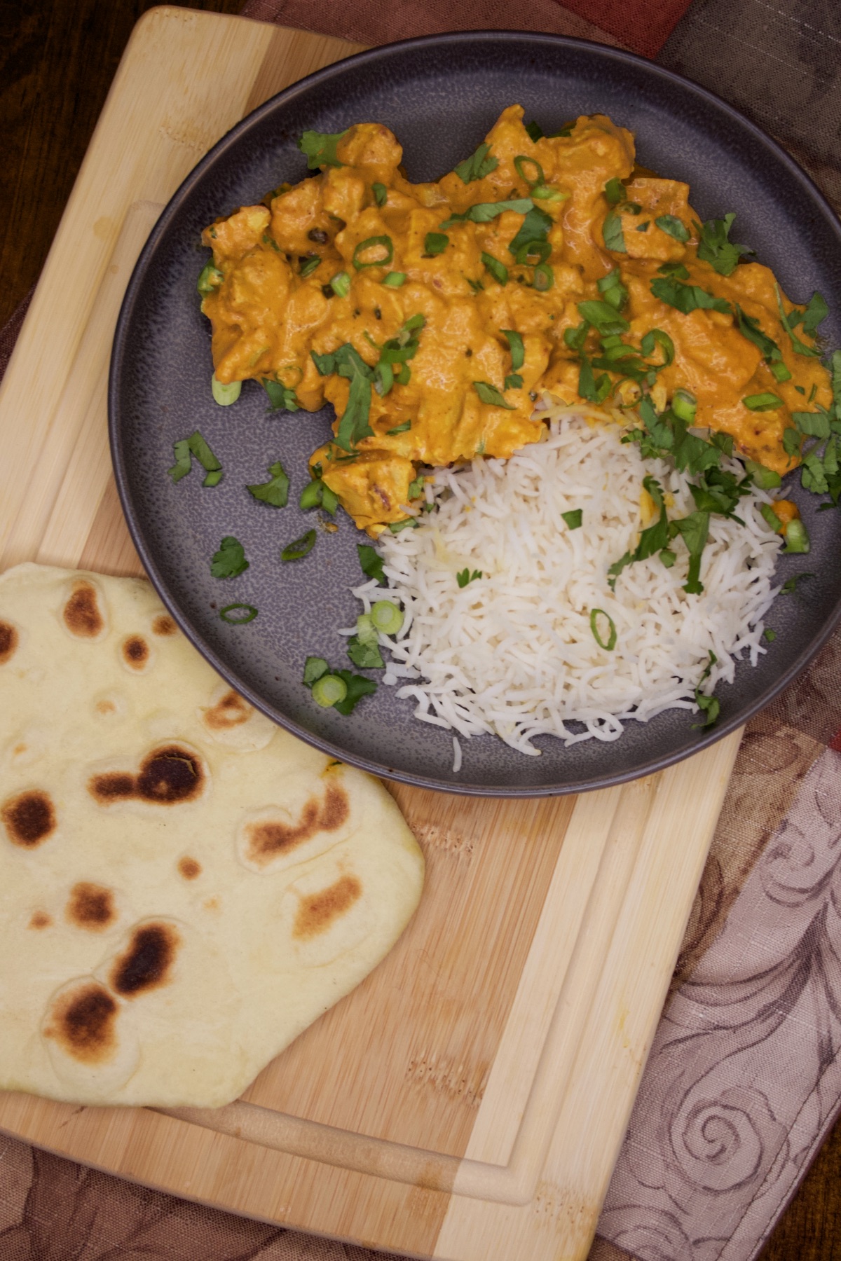 A serving of butter chicken, garnished with chopped cilantro and sliced onions, served with jasmine rice and naan.