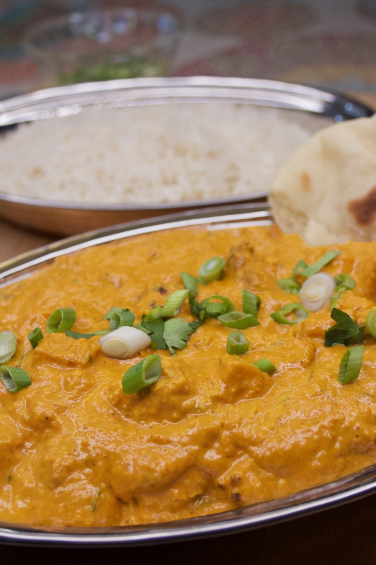 Close-up of butter chicken sprinkled with sliced scallions. A dish of jasmine rice sits in the background.