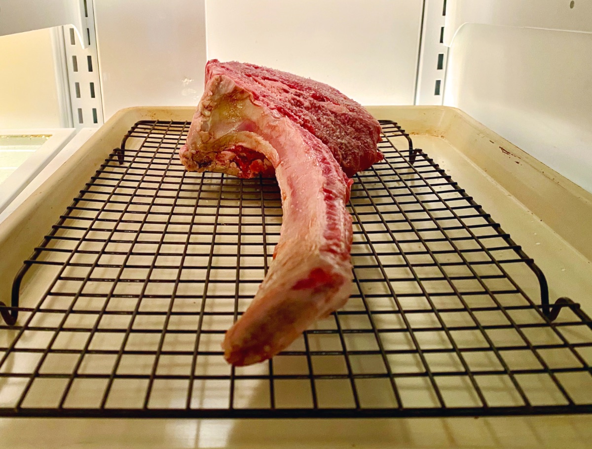 The salted tomahawk steak in the refrigerator for a dry brine. For good air circulation, the steak sits on a wire rack placed inside a baking sheet to catch any drips.