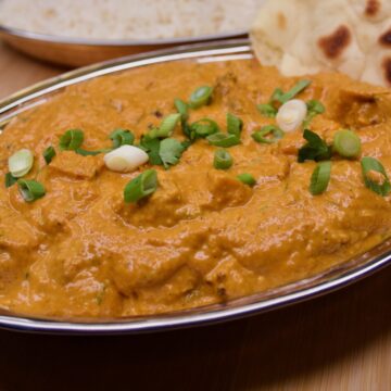 bowl of butter chicken in metal dish with naan bread tucked in it.