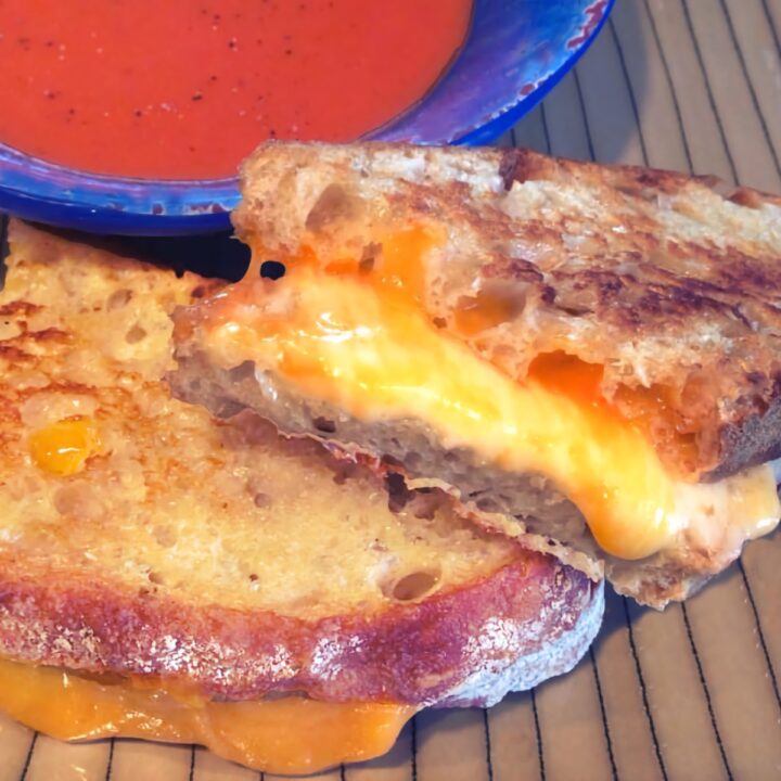 A perfectly gooey grilled cheese sandwich with a bowl of tomato soup in the background.