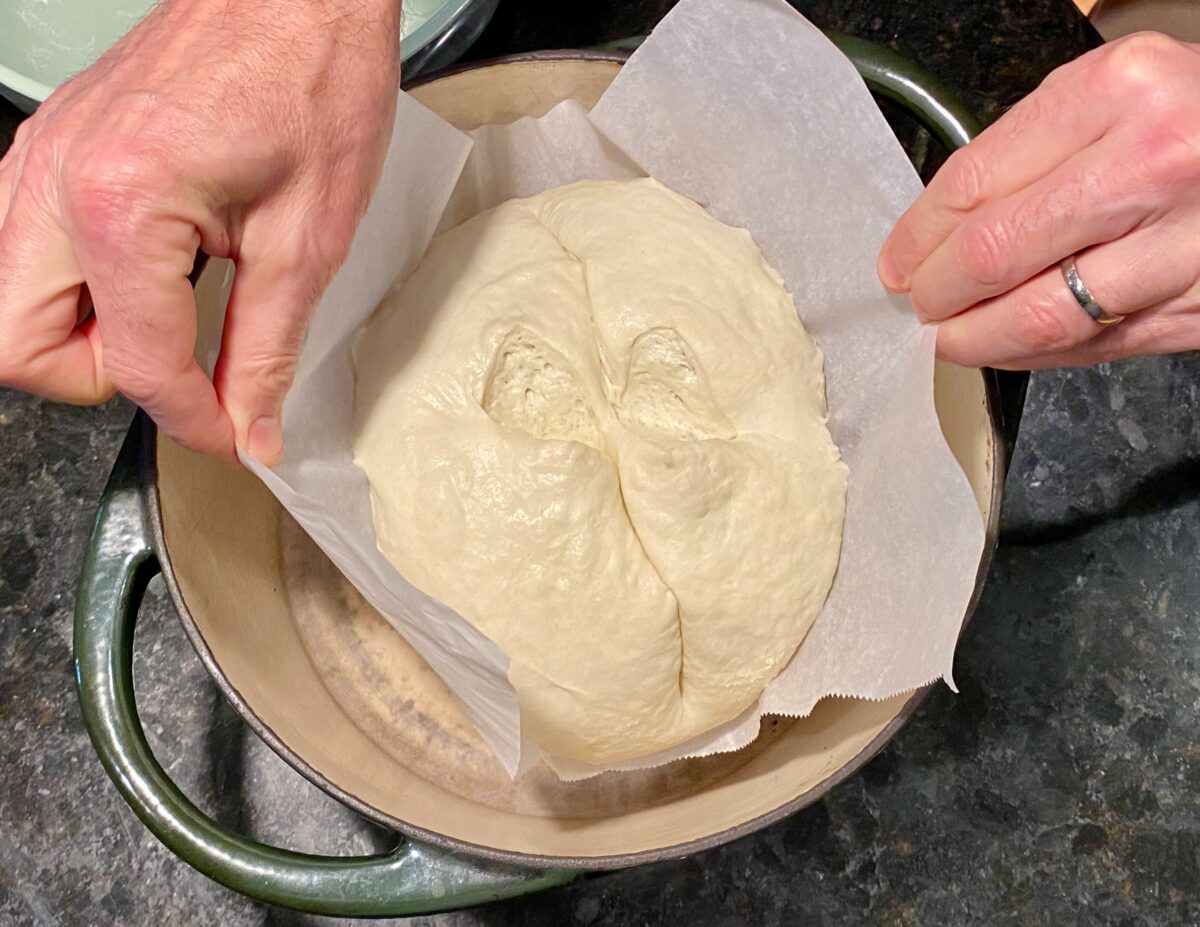 Using the long ends of the parchment paper as handles to carefully lower the dough into the screaming hot Dutch oven.