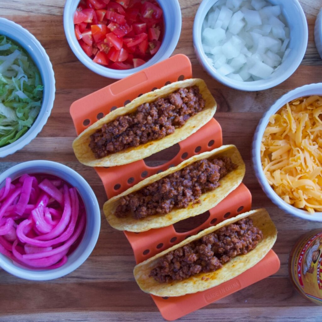 three ground beef tacos in a holder surrounded by toppings.